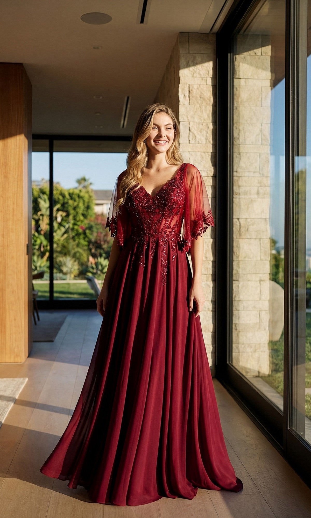 A woman stands indoors in natural light, smiling toward the camera in the Juliet JTM22U Long Formal Dress in burgundy. The elegant gown features a beautiful embroidered sheer bodice with lace details, sheer sleeves, and an a-line floor-length skirt for a flowing formal look.